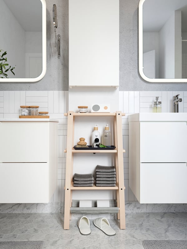 VILTO shelving unit in birch with neatly folded grey towels, a Bluetooth speaker and beauty products on the shelves.