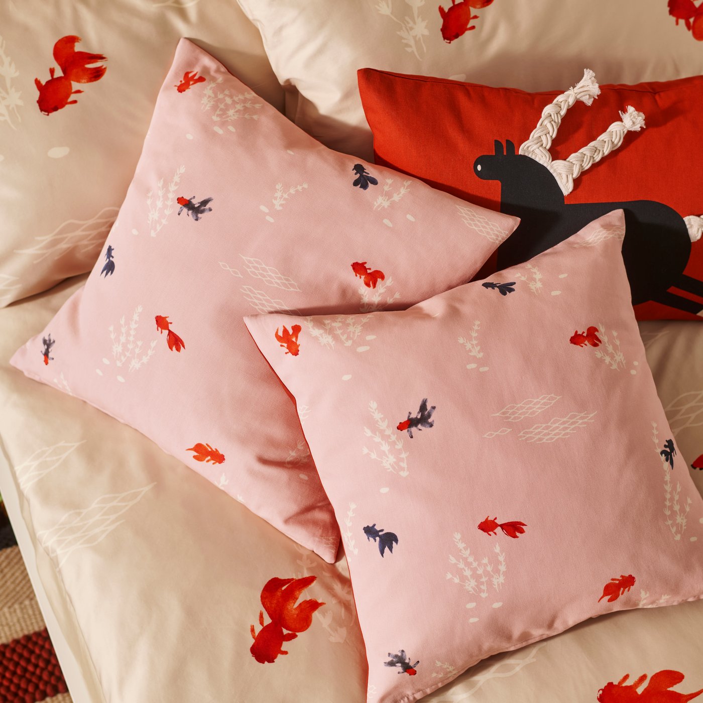 Two FÖSSTA cushion covers with a goldfish design are placed on a bed next to a red FÖSSTA cushion cover with a horse design.