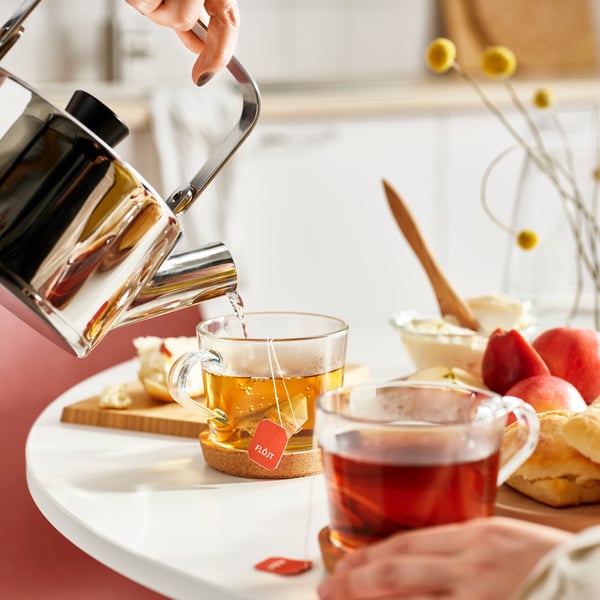 Water is poured from a stainless steel kettle into a clear glass IKEA 365+ mug which has a FLÖJT tea bag in it.