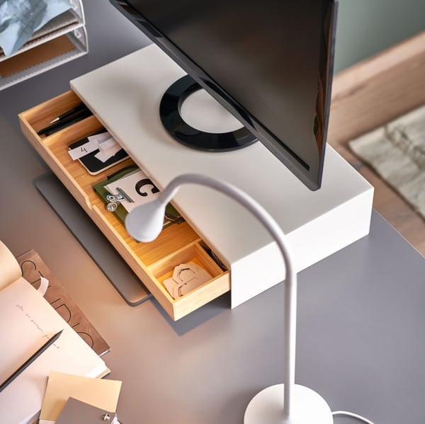 Auf einer grauen Tischplatte steht neben einer ELLOVEN Monitorerhöhung mit Schublade eine NÄVLINGE LED-Arbeitsleuchte in Weiß.