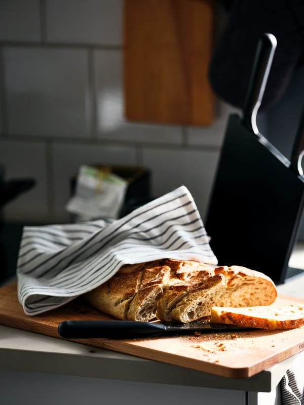 A loaf of bread with a few slices cut from it and a white and dark grey RINNIG tea towel around it on a chopping board.