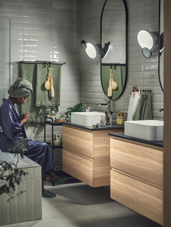Zwei GODMORGON Waschbeckenschränke und zwei LINDBYN Spiegel sind an einer Badezimmerwand zu sehen. Auf der Badewanne sitzt eine Person.