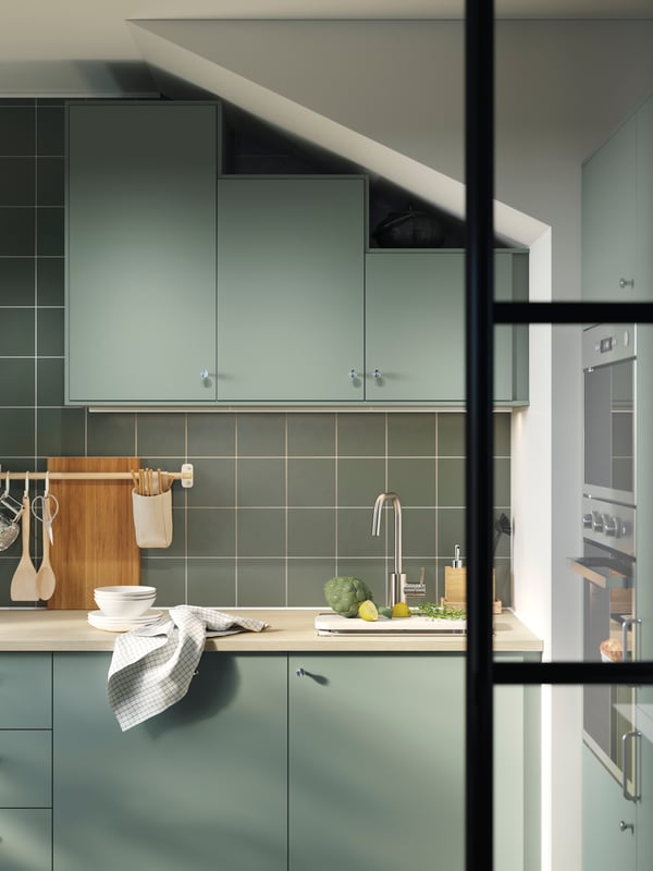 A kitchen has cabinets with grey green NICKEBO fronts and the wall cabinets in different heights follow the slanted ceiling.