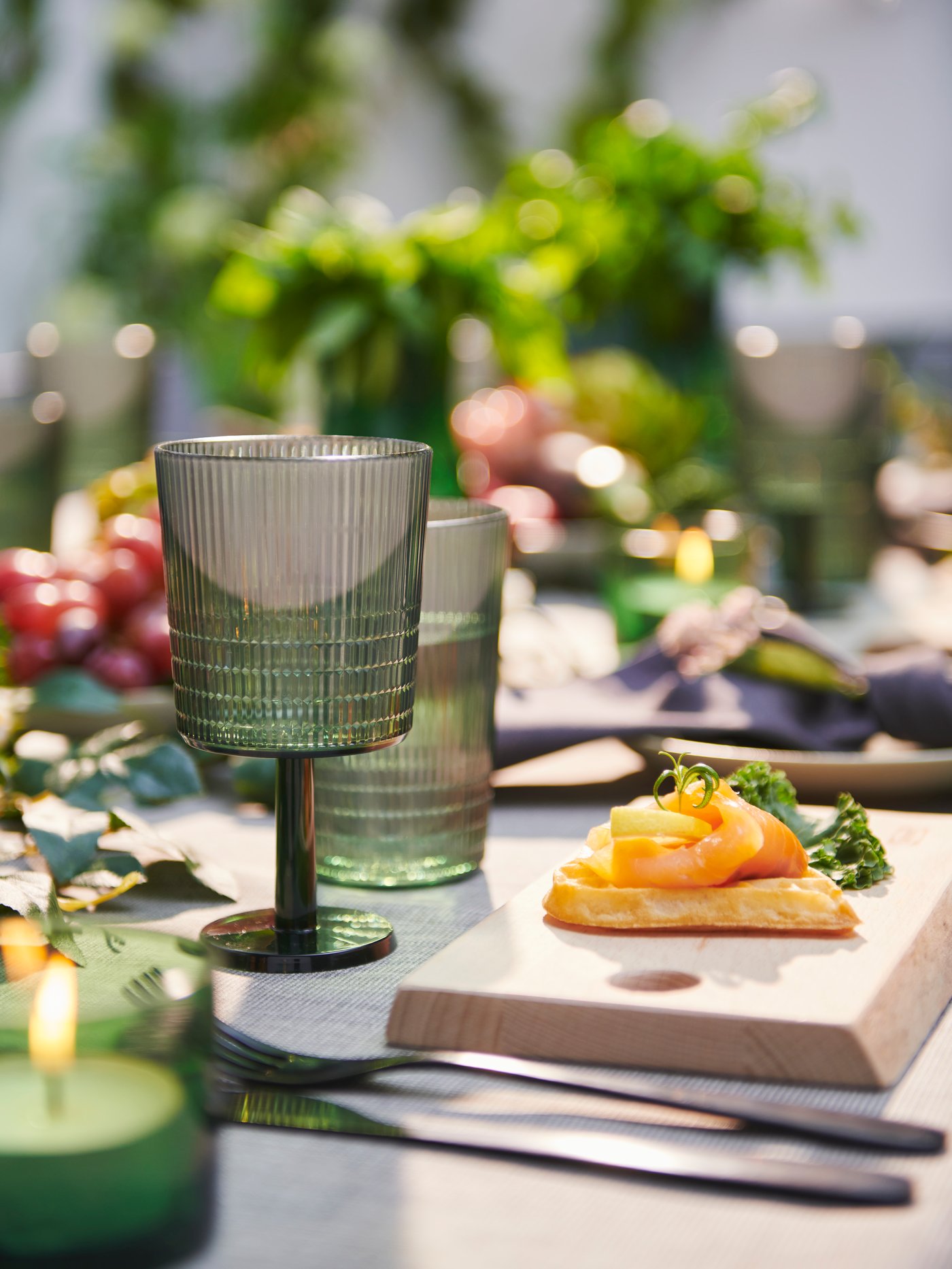 A table is set with a green wine glass and tumbler, cutlery and a wooden cutting board with a Swedish starter on top.