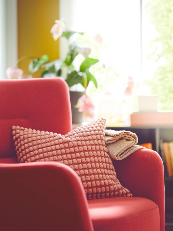 A SVARTPOPPEL cushion cover sits on a red OSKARSHAMN wing chair. In the background a plant sits on a cabinet.