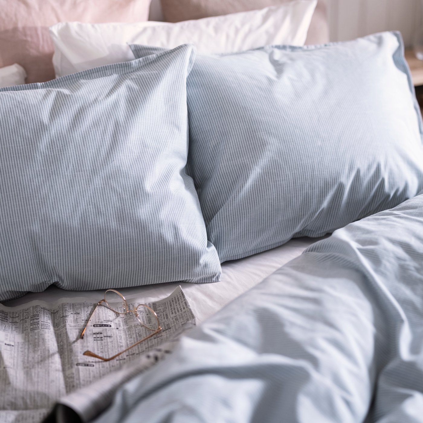 Close-up of a bed with two light blue pillows, one white and one pink pillow, light blue quilt cover and a pair of glasses.