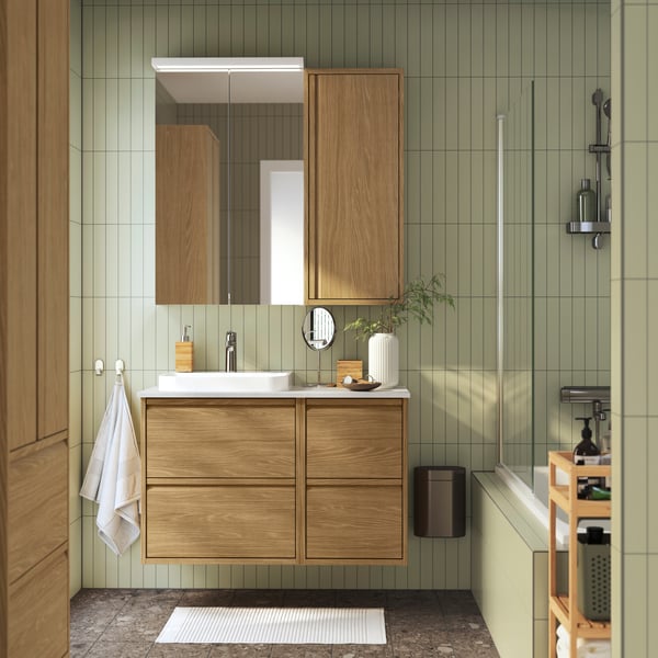 salle de bains ÄNGSJÖN moderne et bien organisée avec des tons de couleur bois, des murs aux carreaux verticaux verts, un meuble avec tiroir ÄNGSJÖN en bois et divers accessoires pour salle de bains.