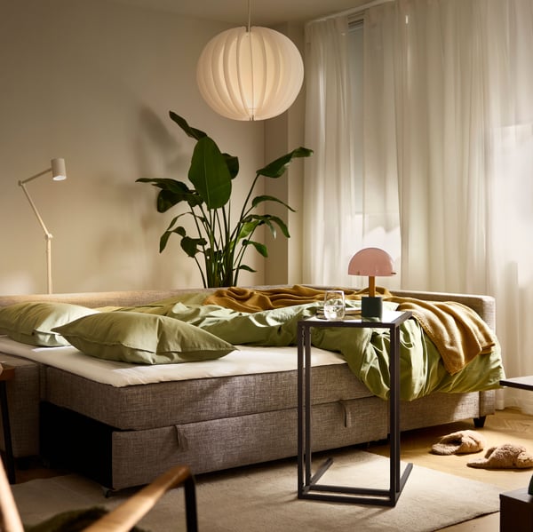 A FRIHETEN/KLAGSHAMN corner sofa-bed, shown as a bed, stands in a softly lit living room with a VOLLERSLEV rug.