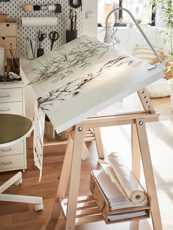 A white angled table top on wooden trestles, dried branches on paper, a white lamp clamped to the table.