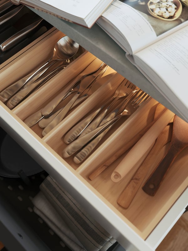 Un cajón abierto que contiene una bandeja de utensilios UPPDATERA de bambú que contiene utensilios BEHAGFULL de acero inoxidable y una cuchara RÖRT.