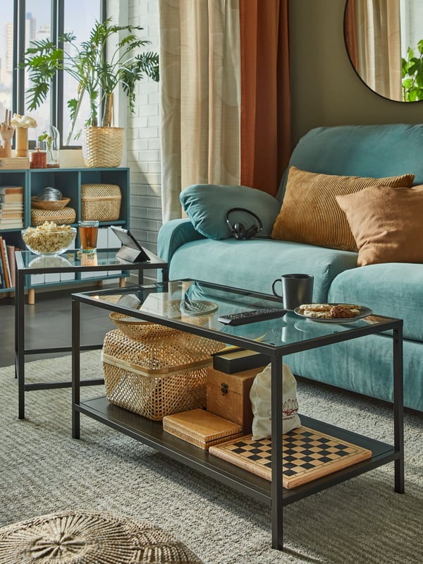 A nest of tables in glass and black metal with games, baskets and boxes below and popcorn, cookies and drinks on top.