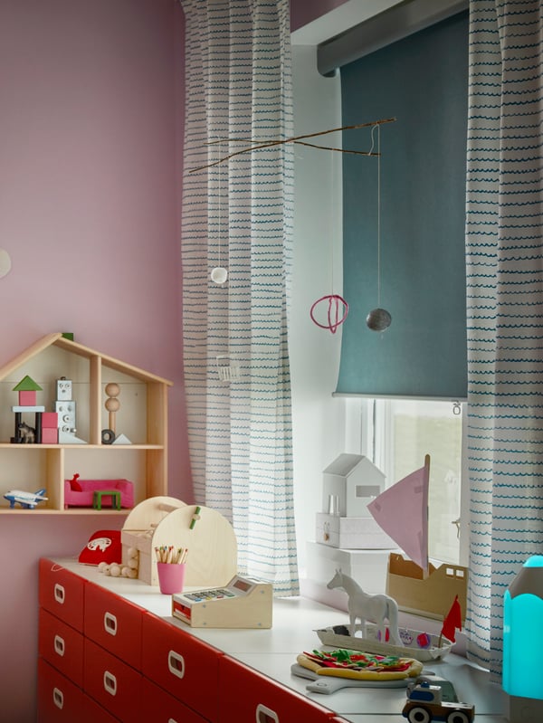 A window in a children’s room with UPPTÅG curtains and a half-closed light blue TRETUR block-out roller blind.