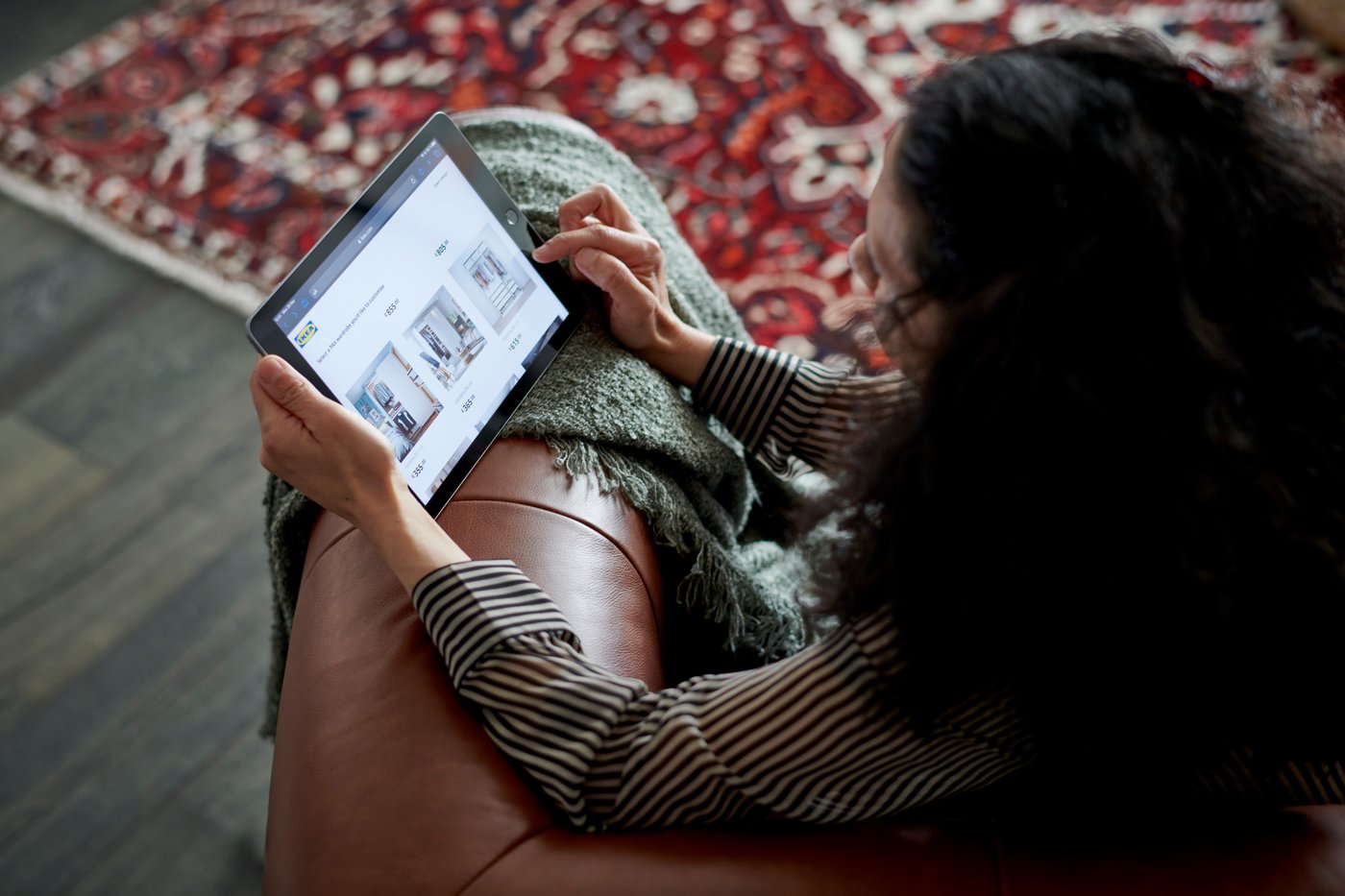A person sits on a brown sofa with a green throw over the arm. They rest a tablet on the arm and browse the IKEA.com website.