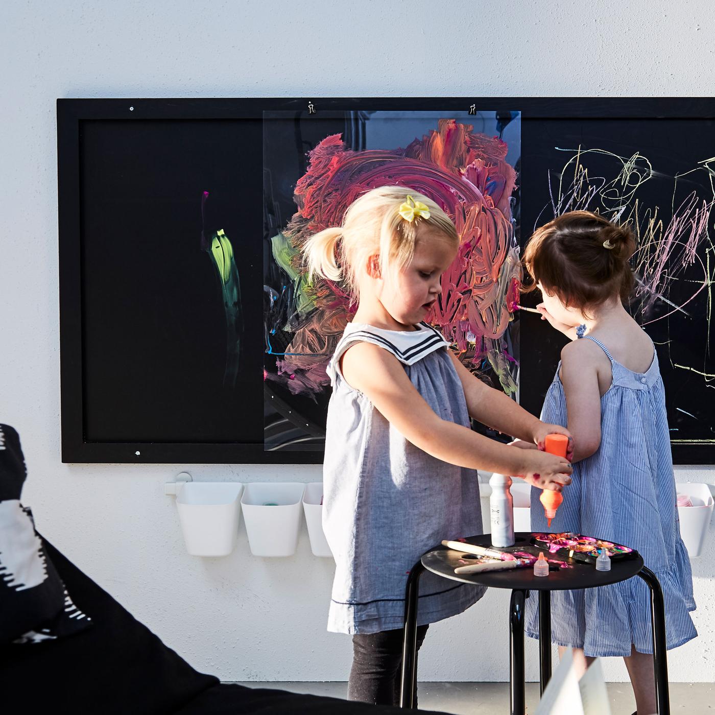 Zwei Kleinkinder drücken Farbe auf Pinsel und spielen mit Kreide. In einer Nahaufnahme ist ein Kindergemälde in einem Kunststoffrahmen zu sehen.