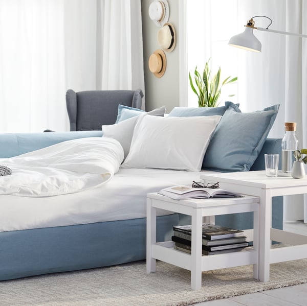 A light room with a light-blue HOLMSUND sofa-bed set up as a bed with pillows and covers, near a white HAVSTA nest of tables.