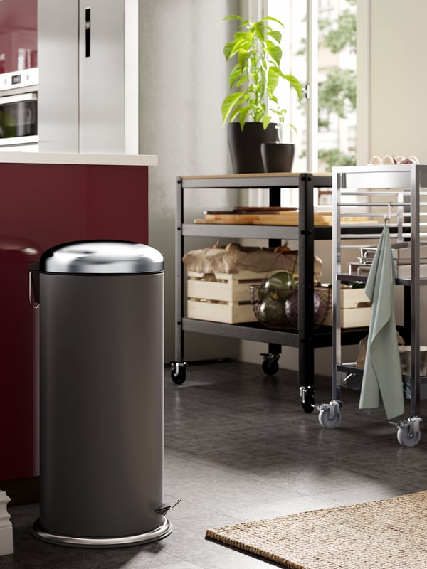 Dark grey MJÖSA pedal bin opposite two kitchen trolleys holding KNAGGLIG pine boxes and plants, in a kitchen with a jute rug.