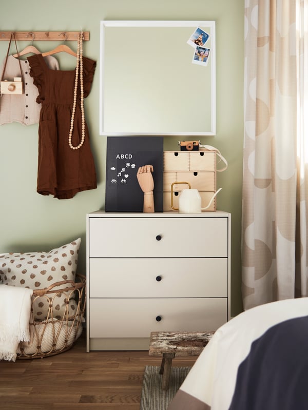 Commode GURSKEN beige clair où sont posés plusieurs articles, surmontée d’un miroir blanc, à côté d’un panier avec un coussin.