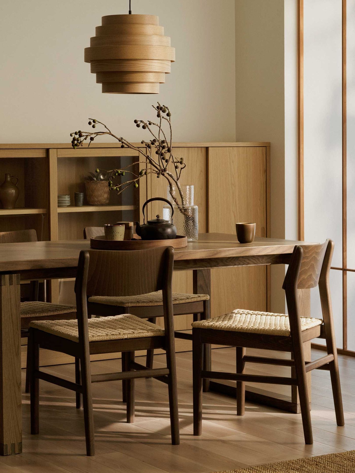 A dining room featuring a long, rectangular wooden dining SKANSNÄS table with four matching wooden SKANSNÄS dining chairs that have woven seats. Above the table hangs a unique, layered wooden pendant light.