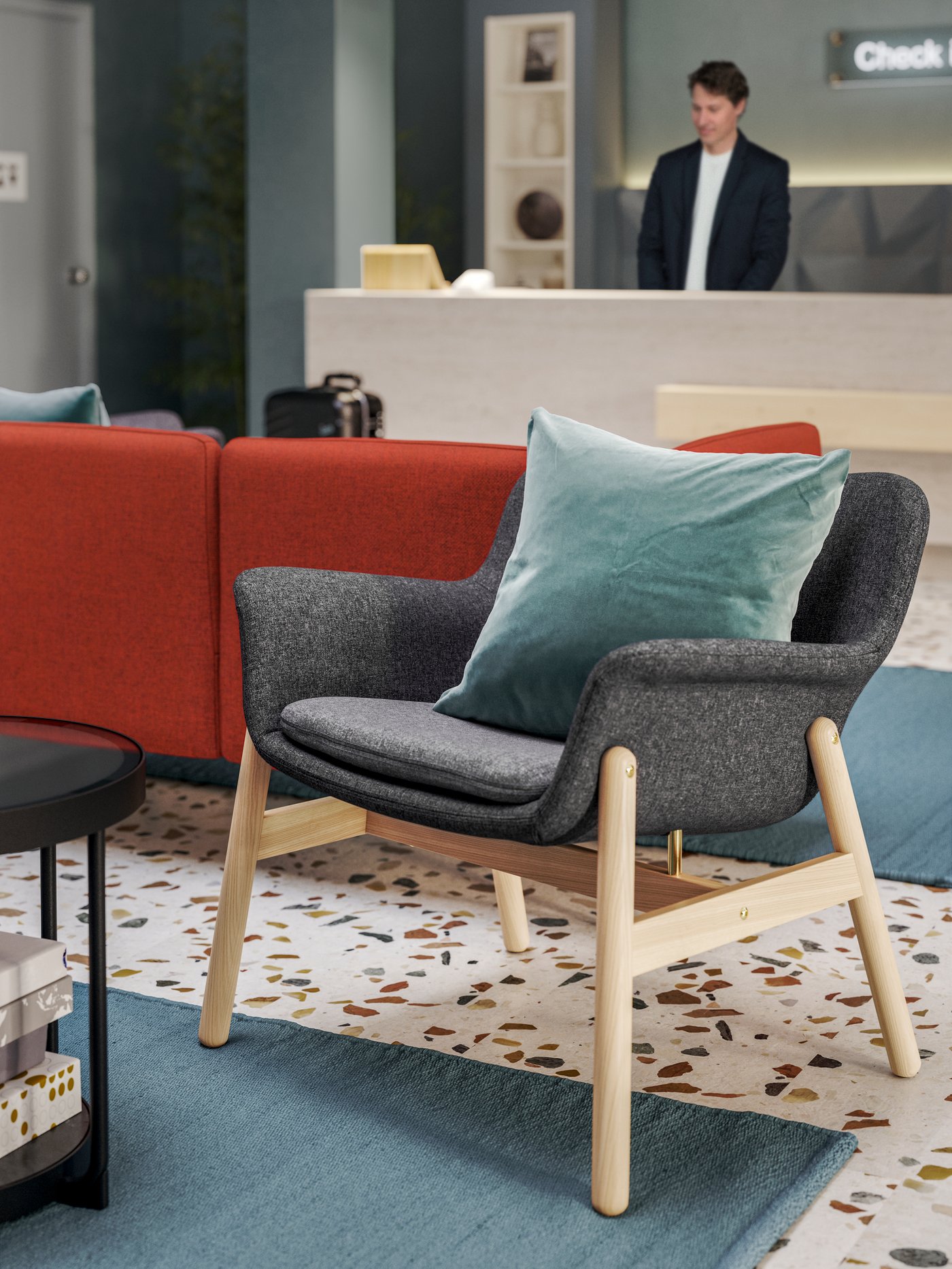 Gegenüber einer Rezepion steht ein dunkelgrauer VEDBO Sessel mit Beinen aus Birke an der Rückseite eines roten Sofas.