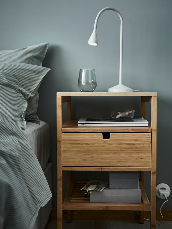 Et sengebord i bambus med en skuffe, står ved siden af sengen og med en lampe oven på.