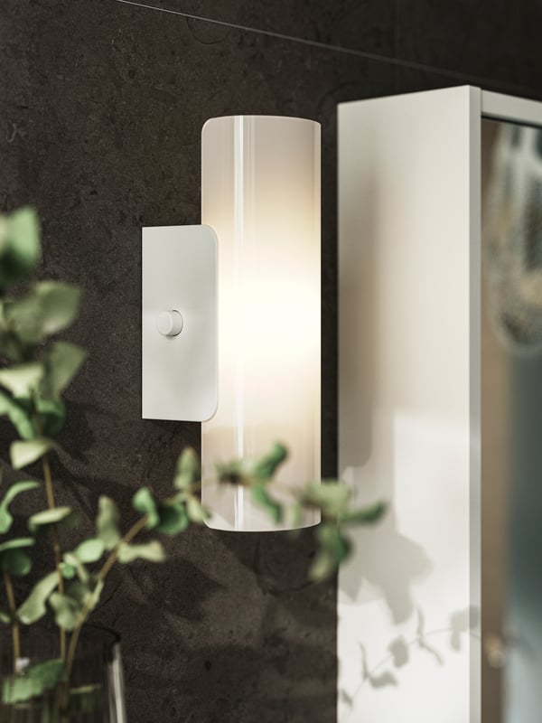 A white HAVSDUN LED wall lamp with frosted glass is dimly lit and hangs next to a mirror on a dark brown wall.