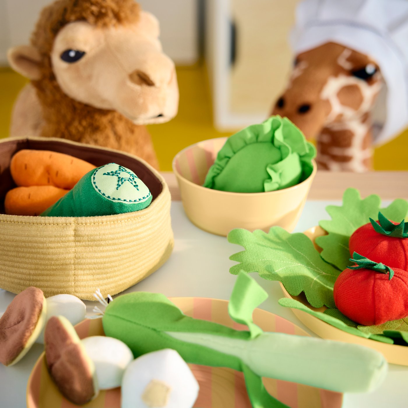 The multicolour LÅTSAS toy vegetables are in a fabric basket and on plates in a child’s room, with a plush camel and giraffe.