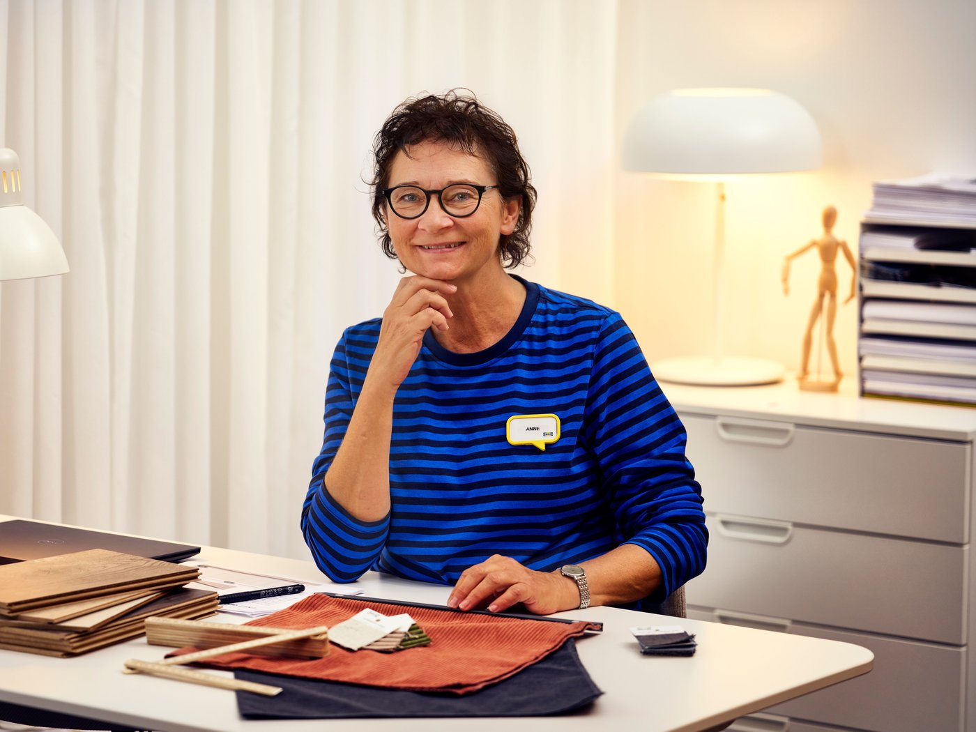 An interior designer from IKEA, wearing a blue sweater, sits at a desk with interior samples and smiles at the camera.