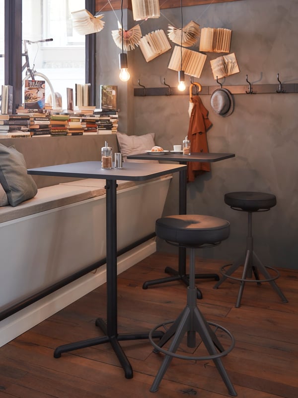 Two dark square tables by a window with books in it, a bench with grey pillows and two black sit and stand supports.