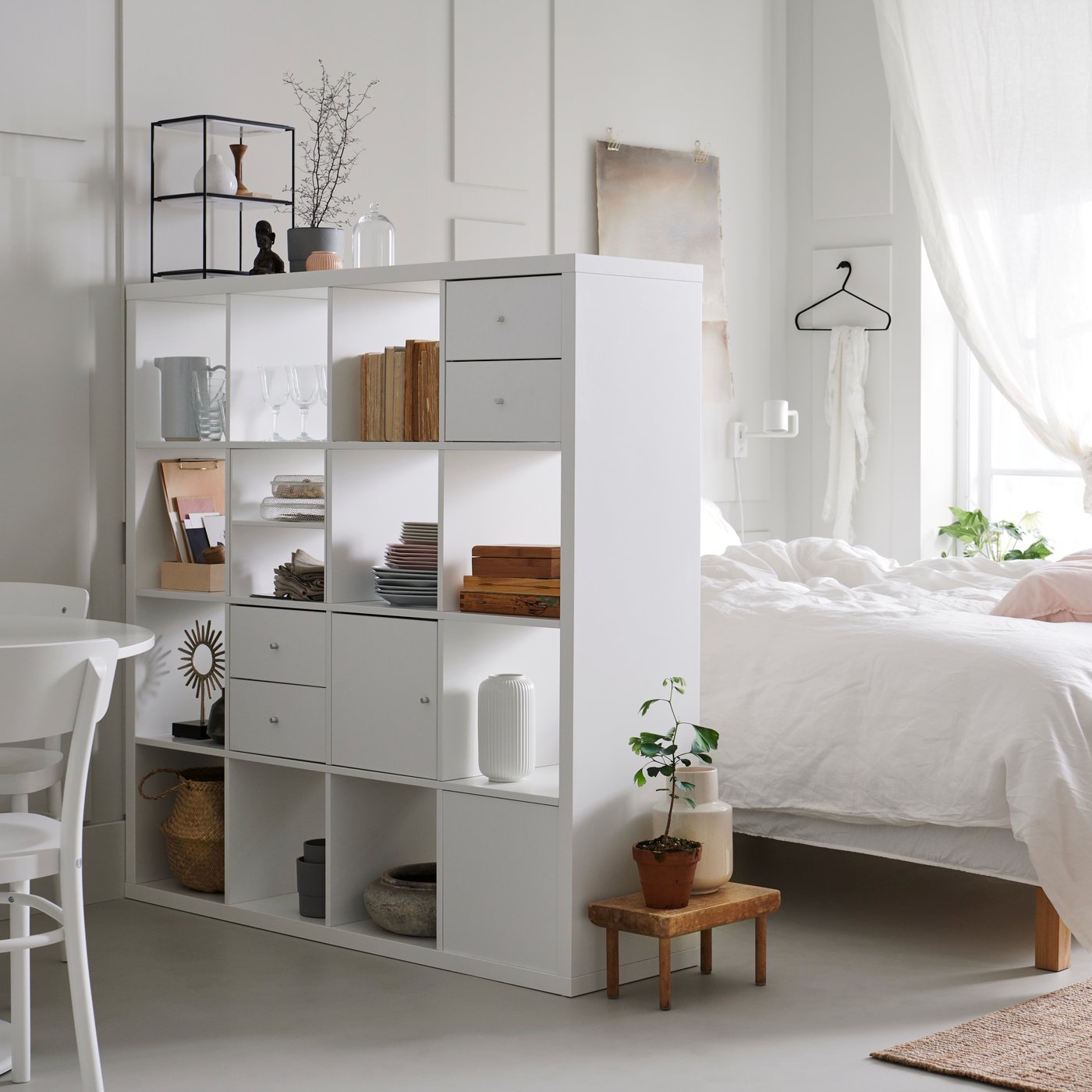 A bedroom featuring a white KALLAX shelving unit filled with decor and storage boxes next to a double bed with white linen.