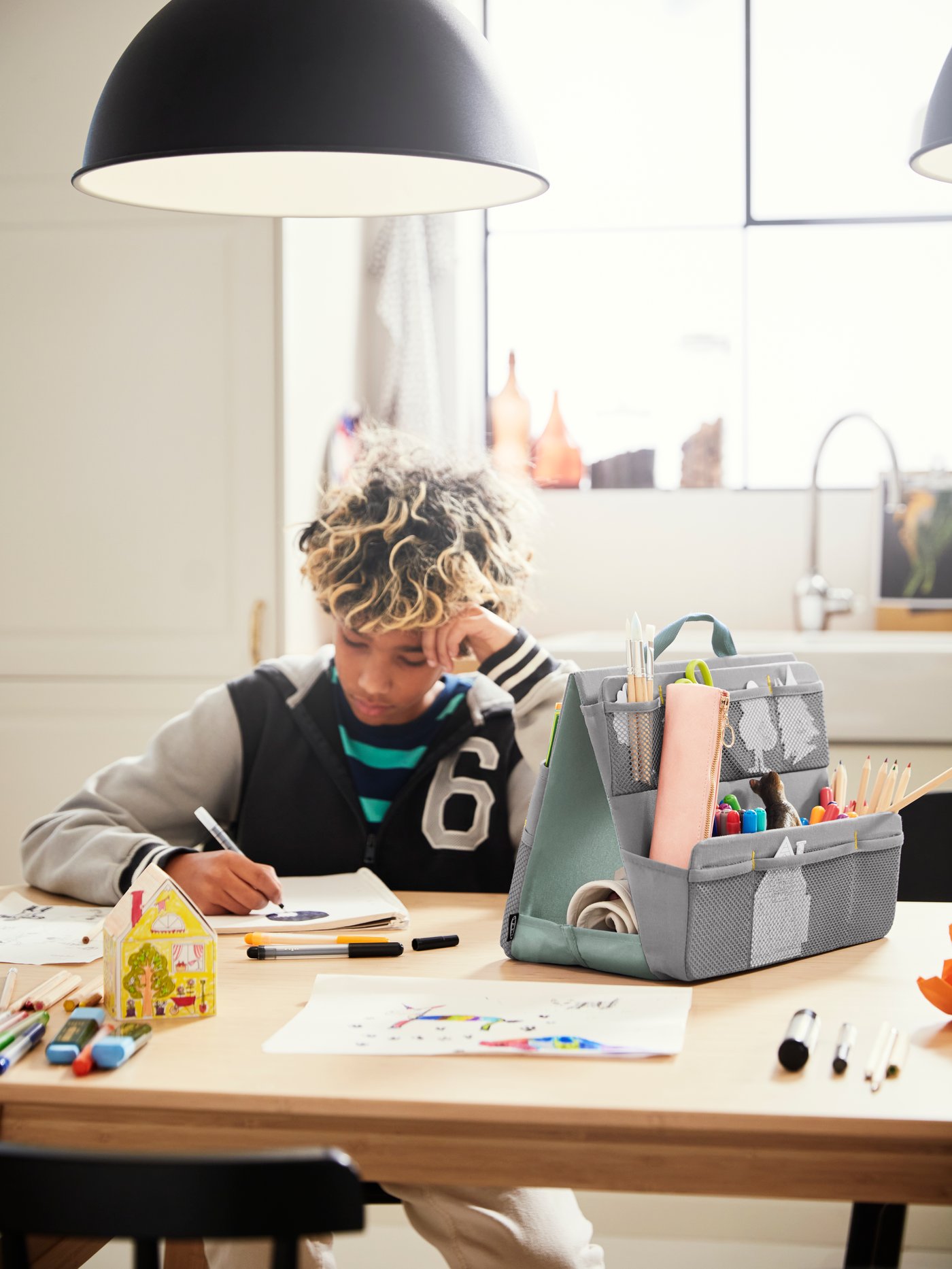 A student sits at a VOXLÖV dining table in light bamboo drawing. An ÖVNING desk accessories organiser sits on the table.