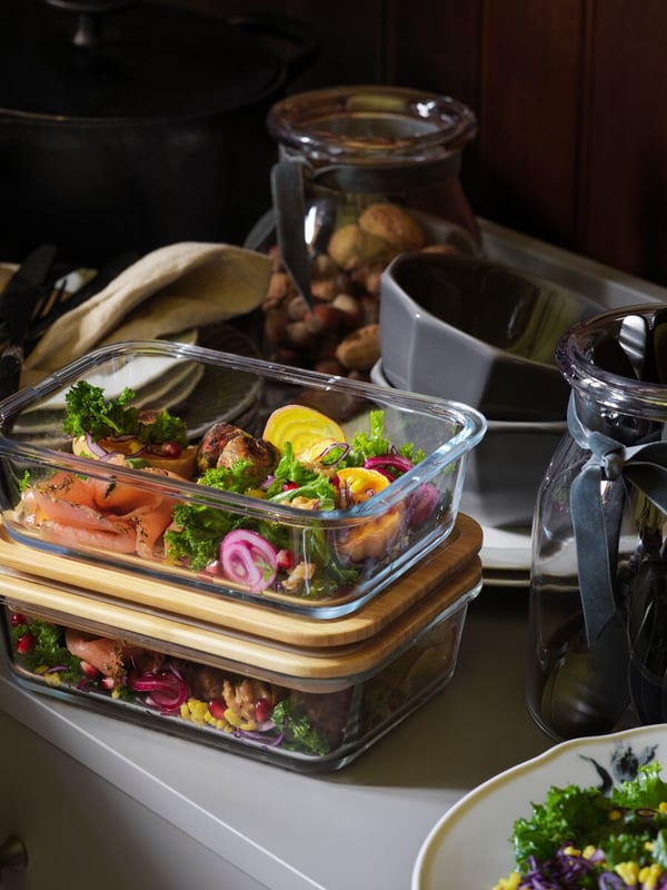 Two rectangular glass containers on top of each other with bamboo lids storing food inside, beside bowls, plates and jars.