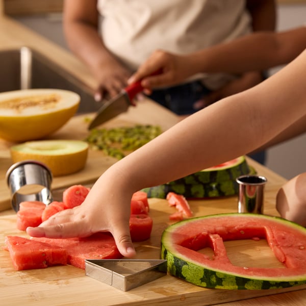 Close up of little children’s hands cutting melon and watermelon pieces with the help of LÄTTBAKAD cookie cutters.