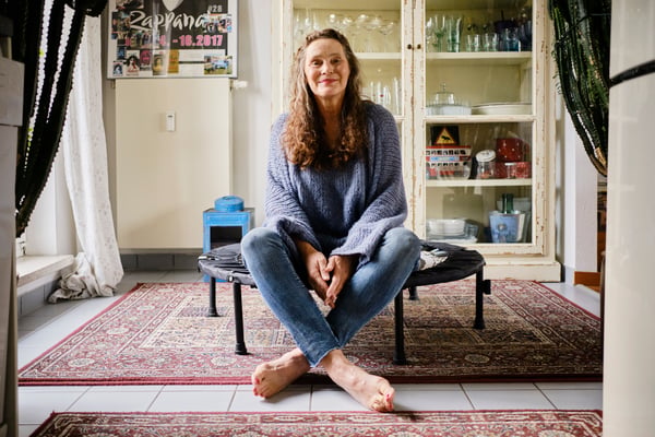 A woman sits on a a mini trampoline, in her living room, smiling and looking at camera. She is surrounded by a plant, a display cabinet and posters.