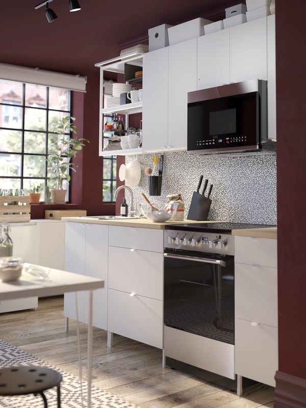 A white ENHET kitchen combination in a deep red room with a desk in front of it. A microwave is above the CIRKULERA range.