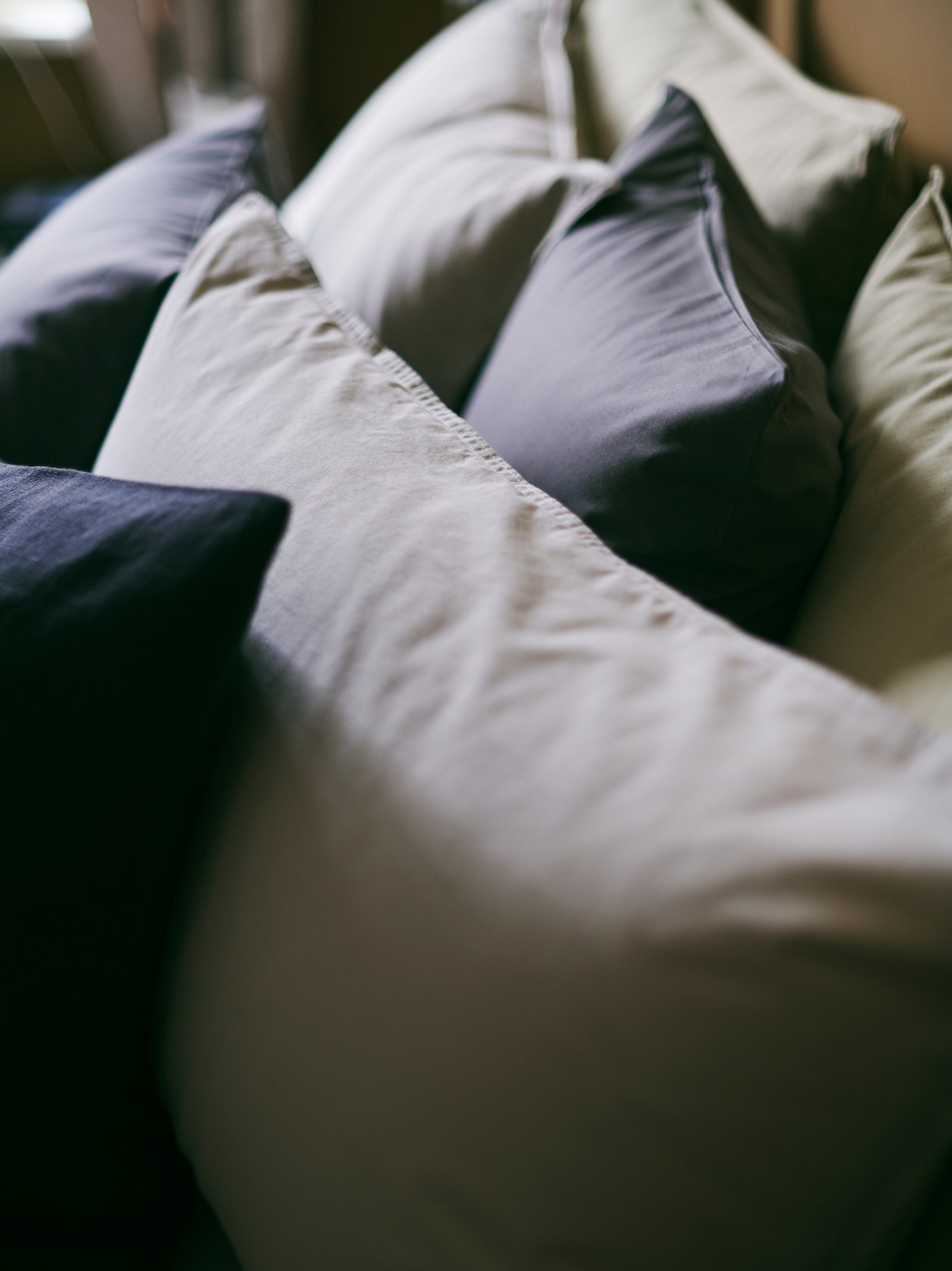 A cosy close-up of ÄNGSLILJA pillowcases in light grey-beige, mixed with dark grey cushions.
