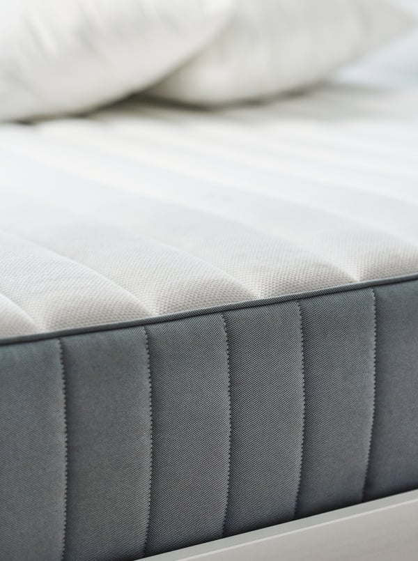 One side of a light blue firm VALEVÅG pocket sprung mattress with a white top surface that has two pillows lying on it.