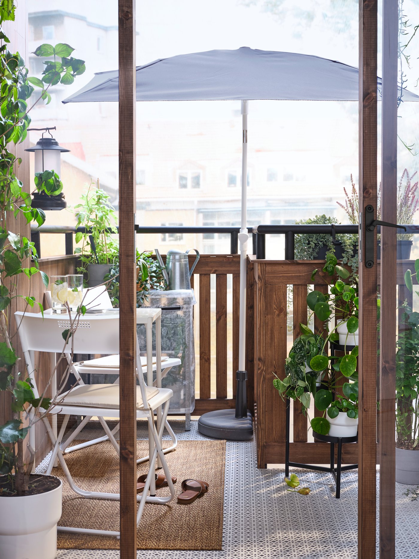 Un petit balcon feuillu avec une table blanche et deux chaises sur un tapis à côté d’un parasol LILLEÖ gris dans un socle BRAMSÖN.