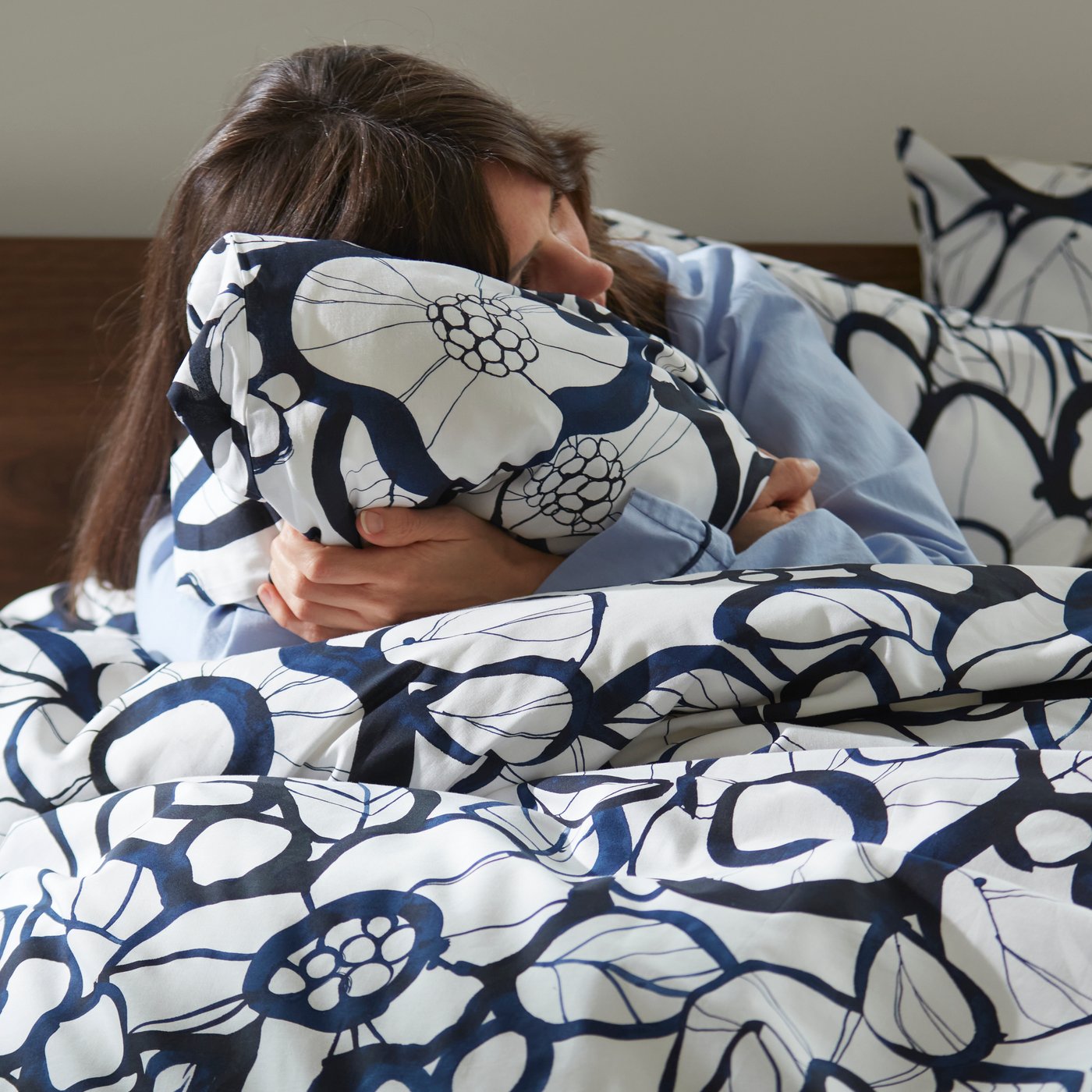 Une personne assise dans un lit serre un oreiller habillé d’une taie HÄRLIR en blanc/noir/bleu, avec une housse de couette assortie.