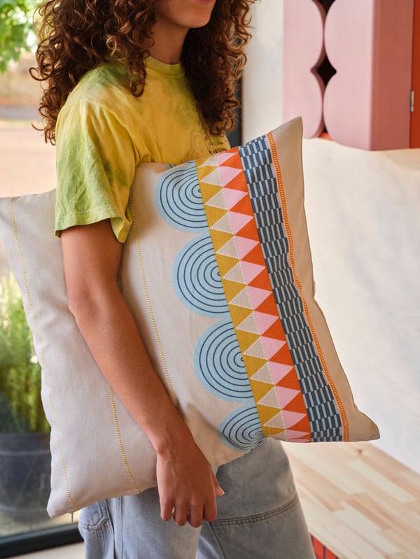 Une personne portant un tee-shirt jaune a sous le bras un coussin habillé d’une housse OMMJÄNGE multicolore.