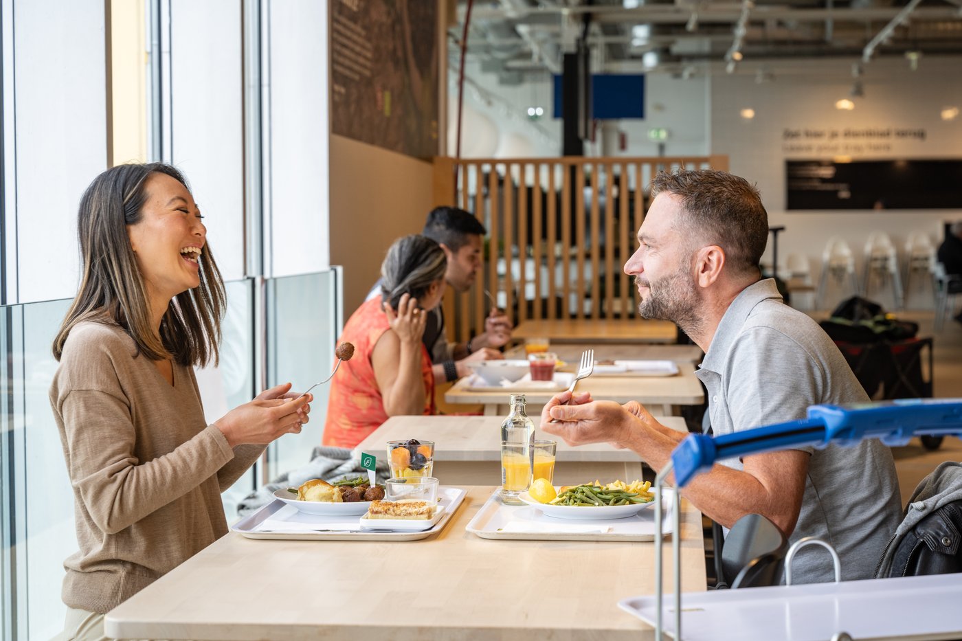Personnes prenant un repas dans un restaurant, avec des plats chauds et des boissons posés sur des plateaux près de la fenêtre.