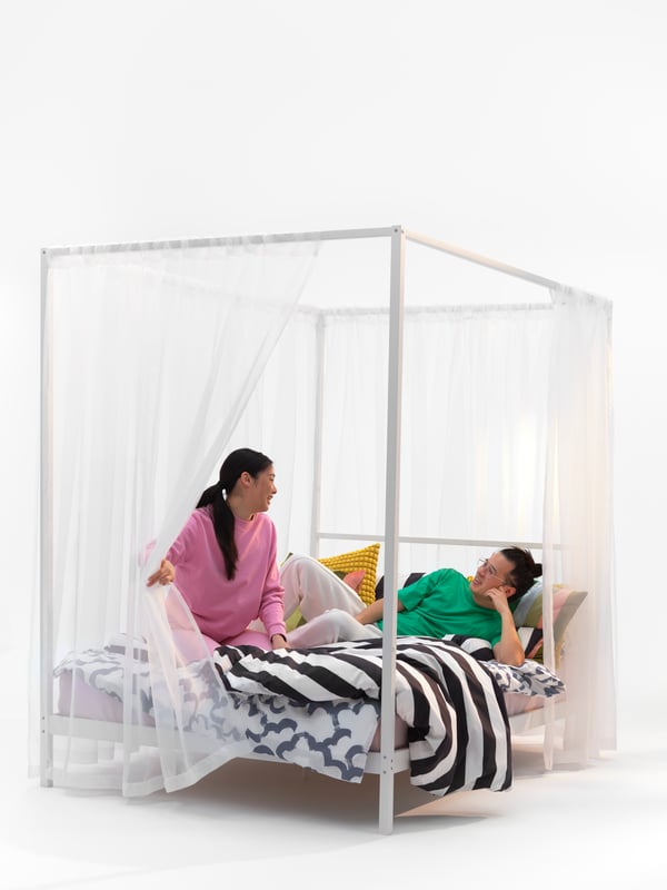 Two people relax in a four-poster bed frame that has gauzy white curtains and cosy bedding.