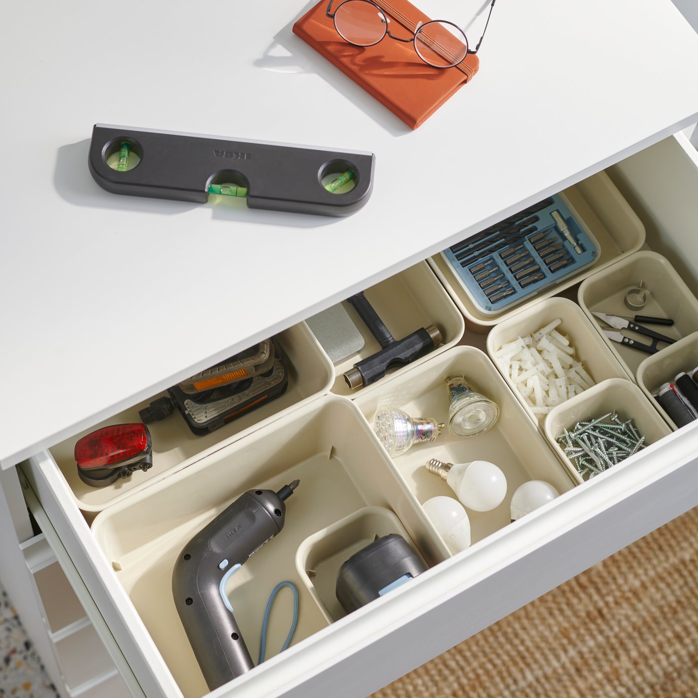 An open drawer of a white STORKLINTA chest of drawers containing organised tools, with a spirit level and glasses on top.