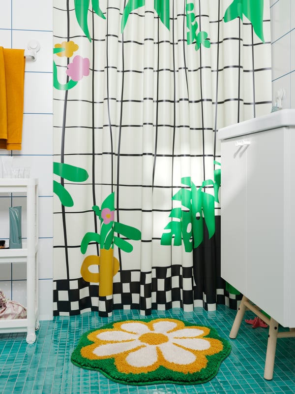 Un piso de baño KÄRRKNIPPROT de flores en amarillo y verde sobre un suelo verde delante de una cortina de baño SNÖJONKVILL en blanco/verde.
