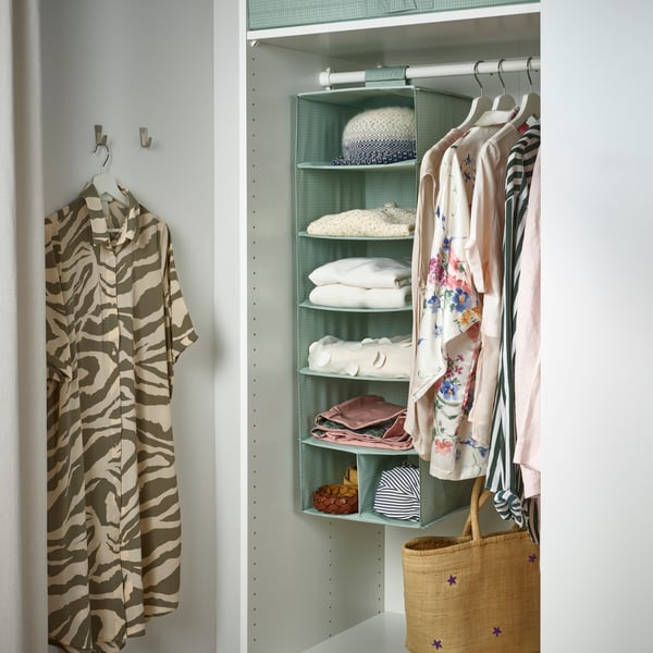 A STUK storage with 7 compartments, holding clothes, headphones, bags and bed linen, hangs from a white MULIG clothes rack.