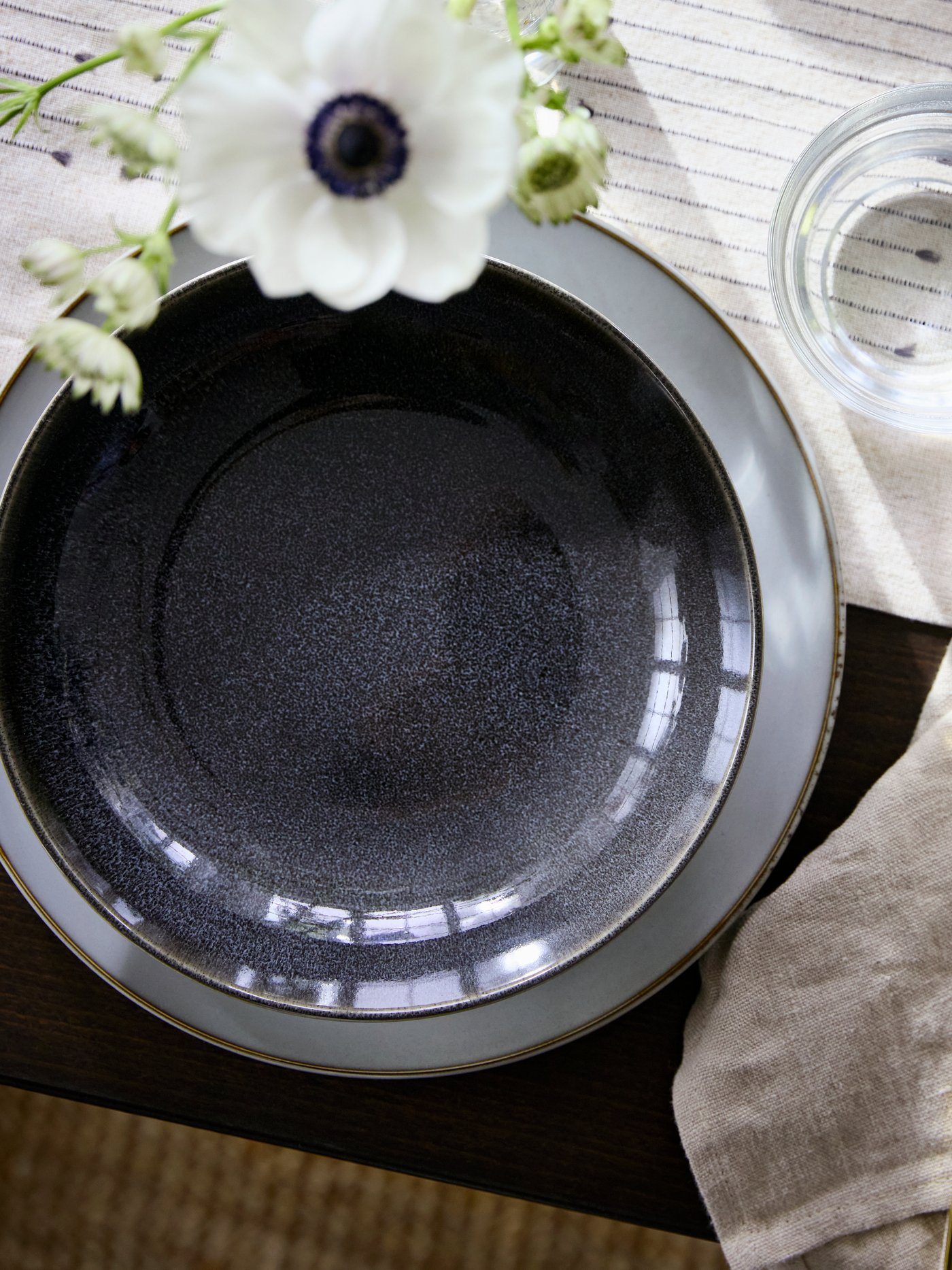 A dark grey GLADELIG deep plate sits on a patterned table with a glass of water, cutlery, and flowers nearby.
