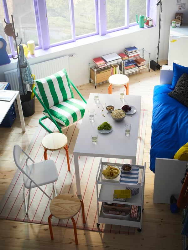 Vue en plongée sur une pièce avec un lit d’appoint et une table entourée de chaises et de tabourets variés. On voit de grandes fenêtres à l’arrière.