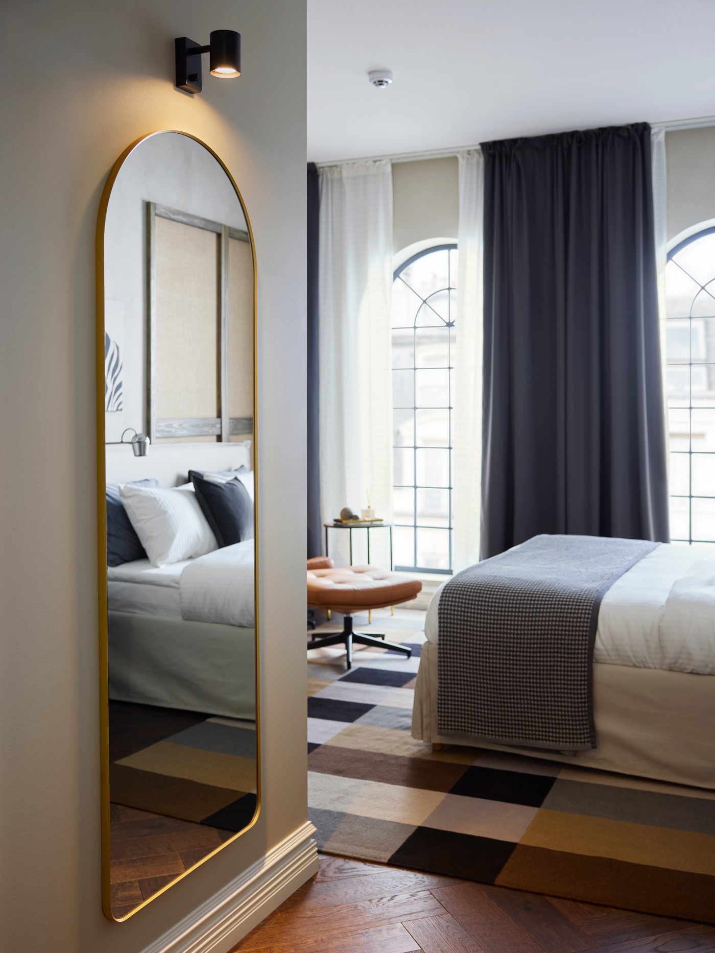 Sotto una lampada da parete illuminata si trova uno specchio LINDBYN con cornice dorata, arrotondato nella parte superiore, che riflette in parte un letto d'albergo.