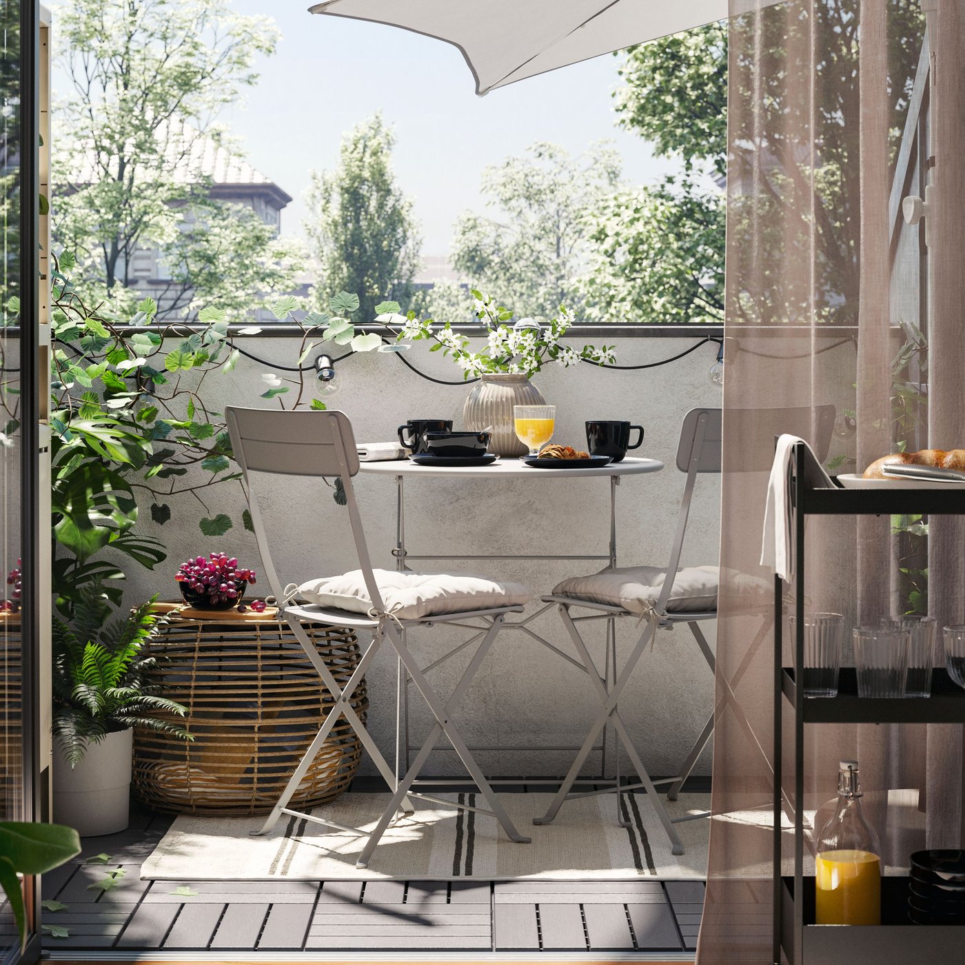 Un balcone con un tavolo e due sedie da giardino SUNDSÖ in grigio su un tappeto, oltre a un tavolino, piante e un ombrellone.