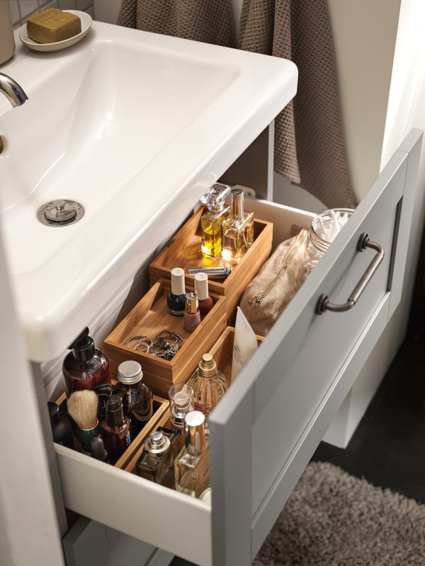 An open drawer with an ENHET drawer front with grey frame, DRAGAN boxes in bamboo with items and a TVÄLLEN washbasin single.