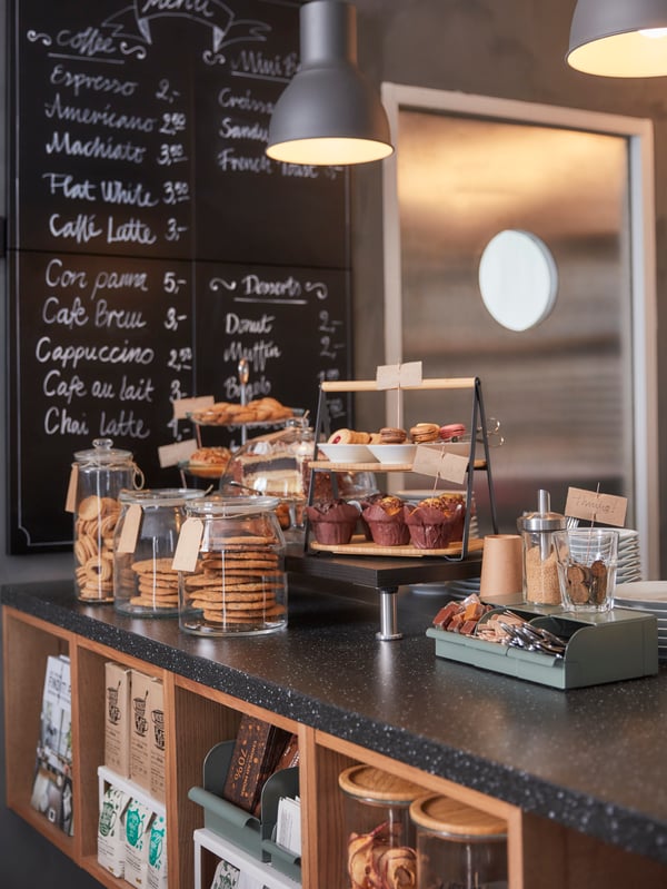 A café kitchen counter with glass jars filled with biscuits, a serving tray in two levels, a green box and white plates.